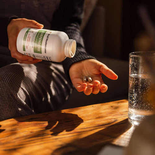 A person pours 5 Defenders® Organic Mushroom Blend Capsules by Real Mushrooms into their hand, showing immune support, with a glass of water on a wooden table nearby.