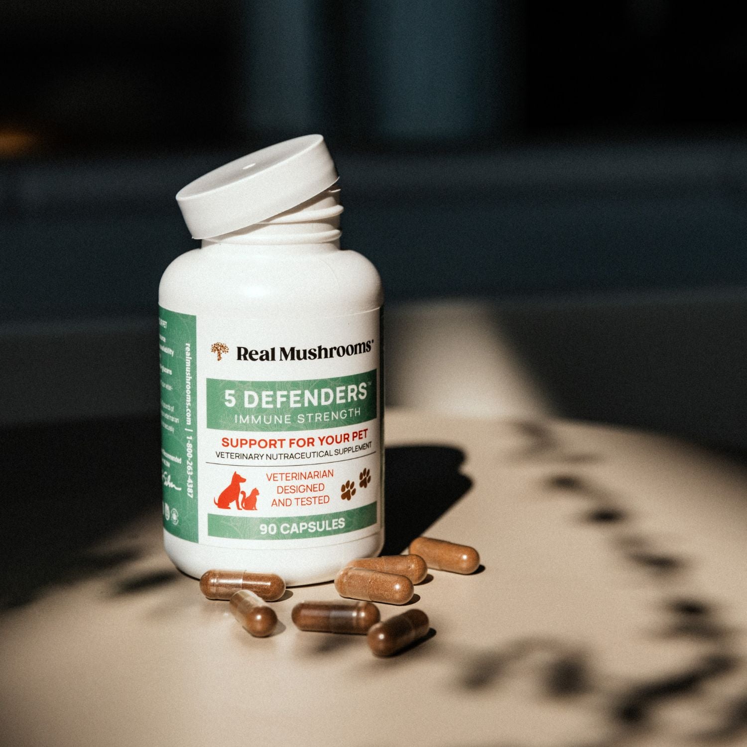 A bottle of Real Mushrooms 5 Defenders® Organic Mushroom Blend Capsules for Pets is displayed on a surface with several capsules beside it.