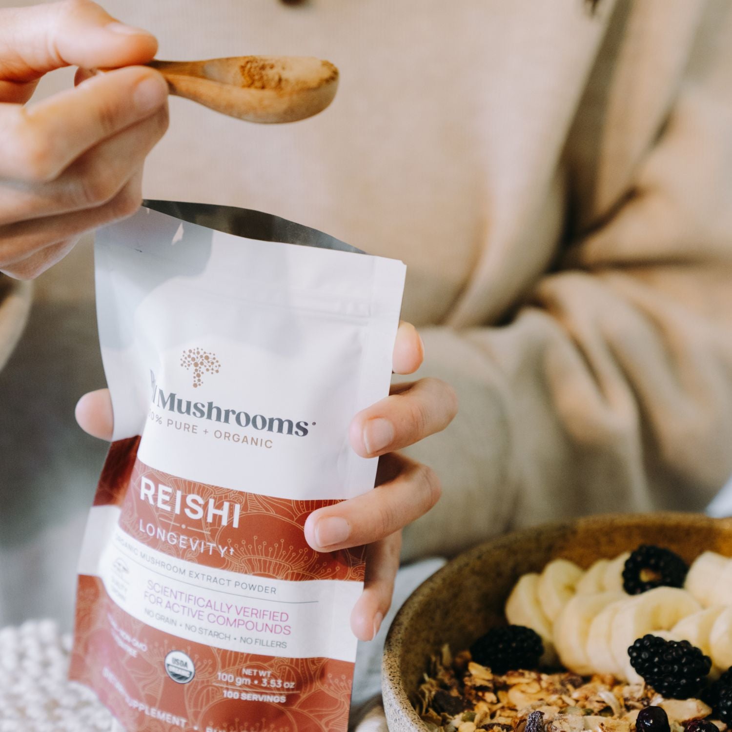 A person scoops Real Mushrooms Organic Reishi Mushroom Powder – Bulk Extract from a bag over a bowl of granola with blackberries, adding an organic reishi supplement rich in beta-glucans to their breakfast.