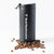 A black 12oz Travel Tumbler from Real Mushrooms stands upright with its lid open, surrounded by scattered coffee beans on a white background.