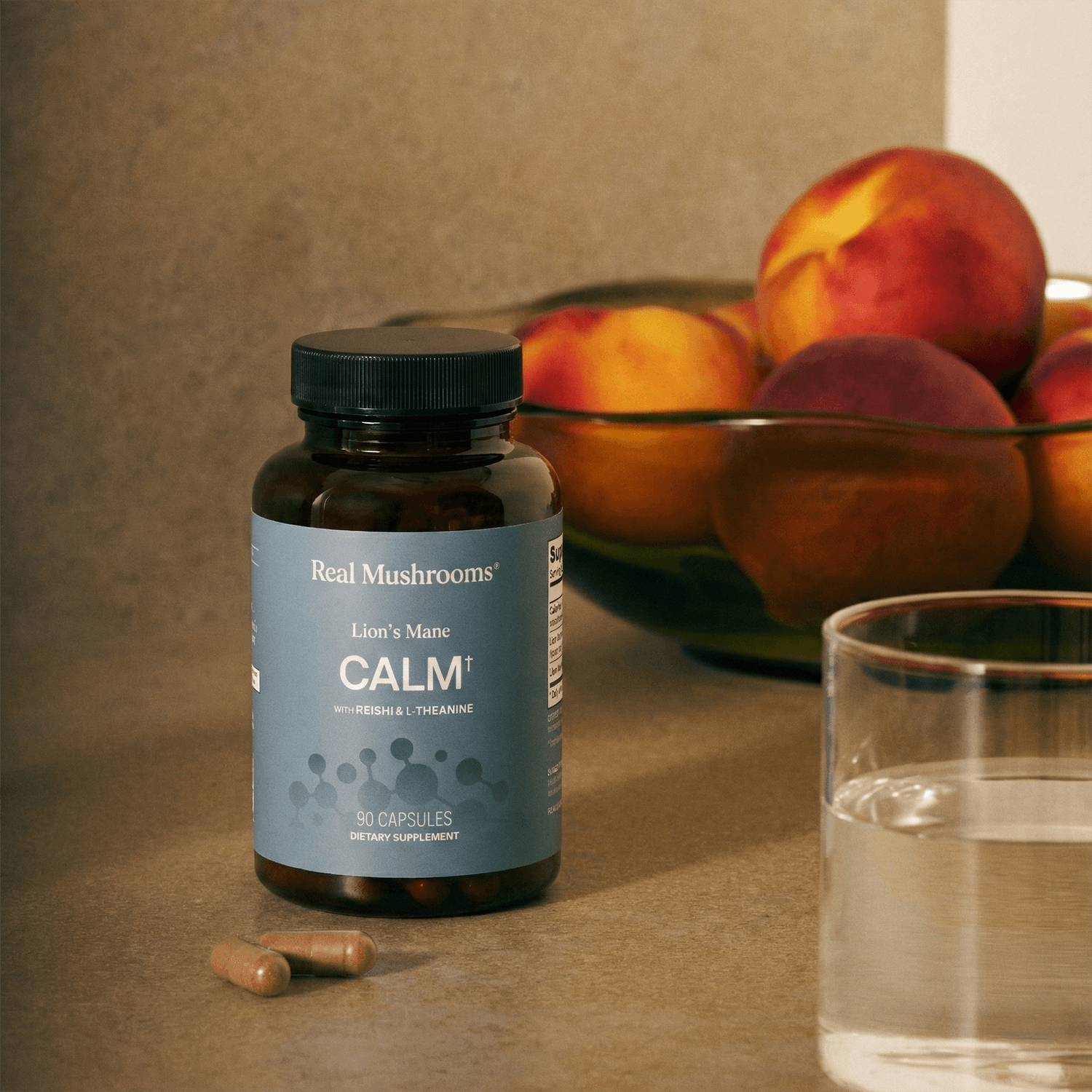 A bottle of Real Mushrooms Lion's Mane Calm sits on a counter beside two capsules, a glass of water, and a bowl of peaches.