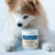 A fluffy dog looks down at a container of Real Mushrooms Daily Dawg™ Powder for Pets, an organic supplement placed in front of its paws.