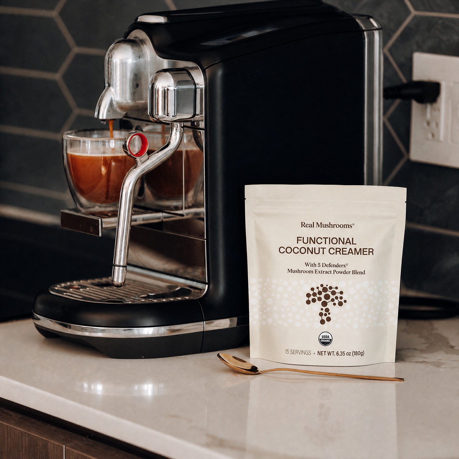A coffee machine brews espresso on a countertop beside a gold spoon and a bag of Real Mushrooms Functional Coconut Creamer - Powder.