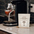 A coffee machine brews espresso on a countertop beside a gold spoon and a bag of Real Mushrooms Functional Coconut Creamer - Powder.