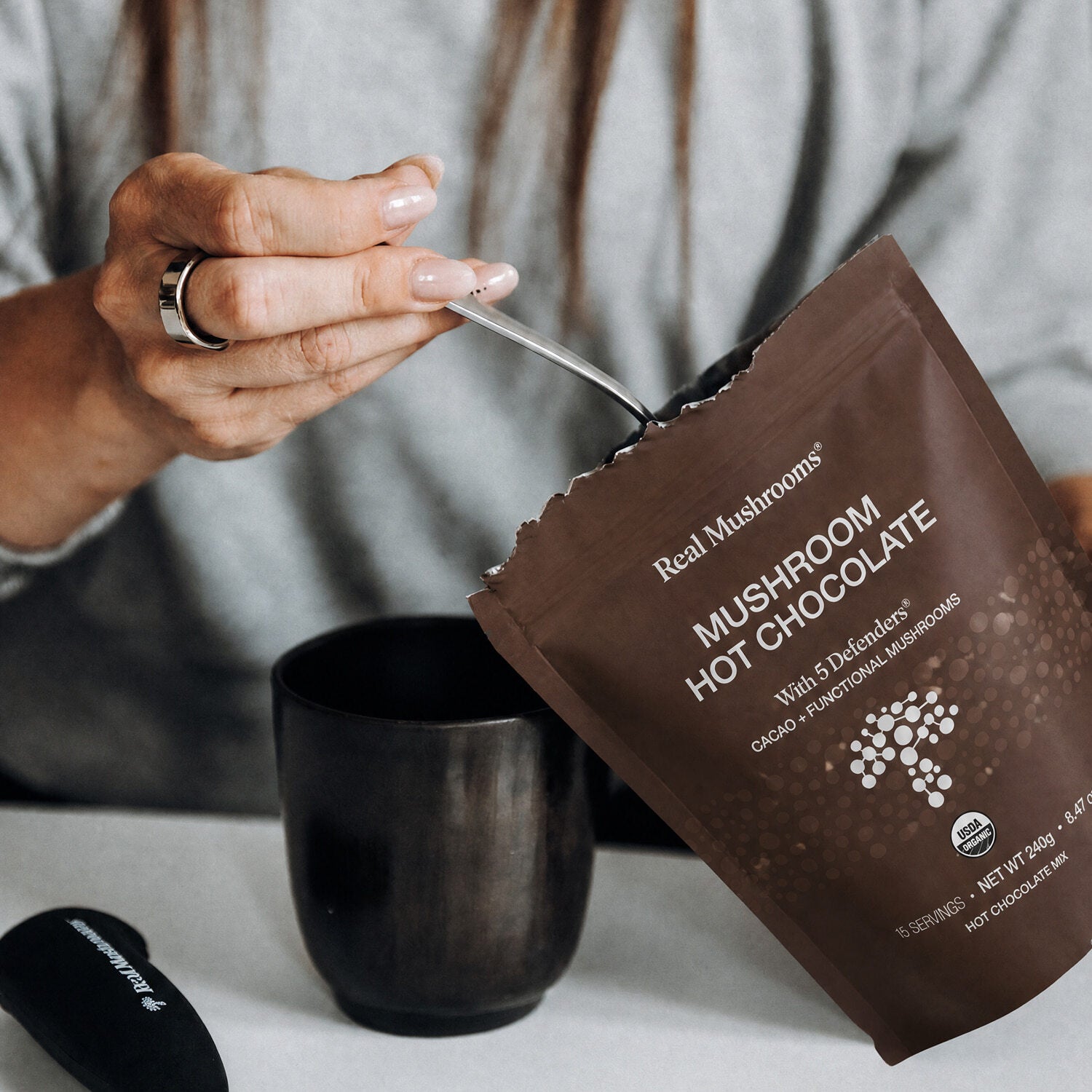 Someone scoops Real Mushrooms Mushroom Hot Chocolate Mix from a brown packet into a black mug, preparing a cozy vegan hot chocolate.