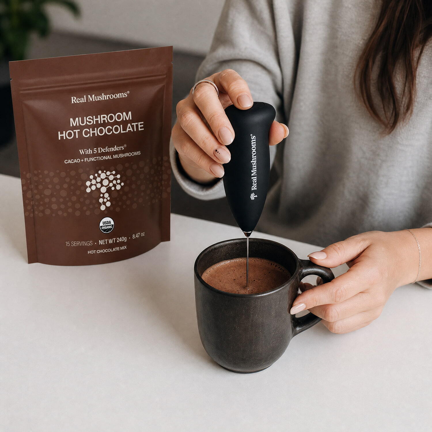 Someone uses a handheld frother to mix vegan hot chocolate in a mug, with a bag of Real Mushrooms Mushroom Hot Chocolate Mix on the table.