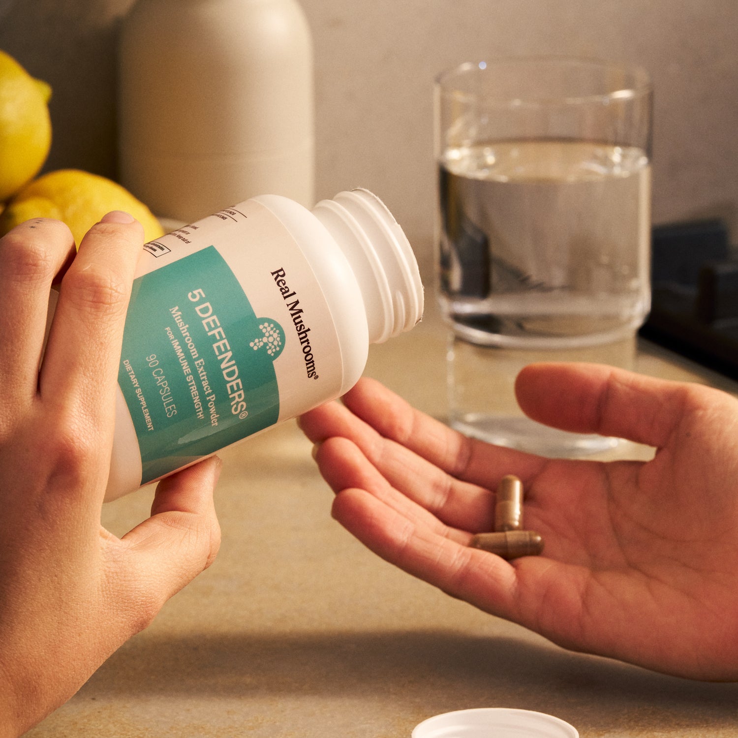 A person pours Real Mushrooms 5 Defenders® Organic Mushroom Blend Capsules into another’s hand, with a glass of water and lemons in the background, emphasizing natural immune support.