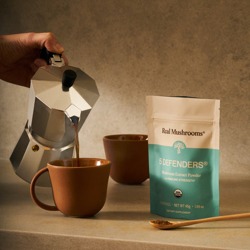 A hand pours coffee from a moka pot into a brown mug beside a packet of Real Mushrooms 5 Defenders® Organic Mushroom Complex – Bulk Powder and a wooden spoon with powder on a beige countertop.
