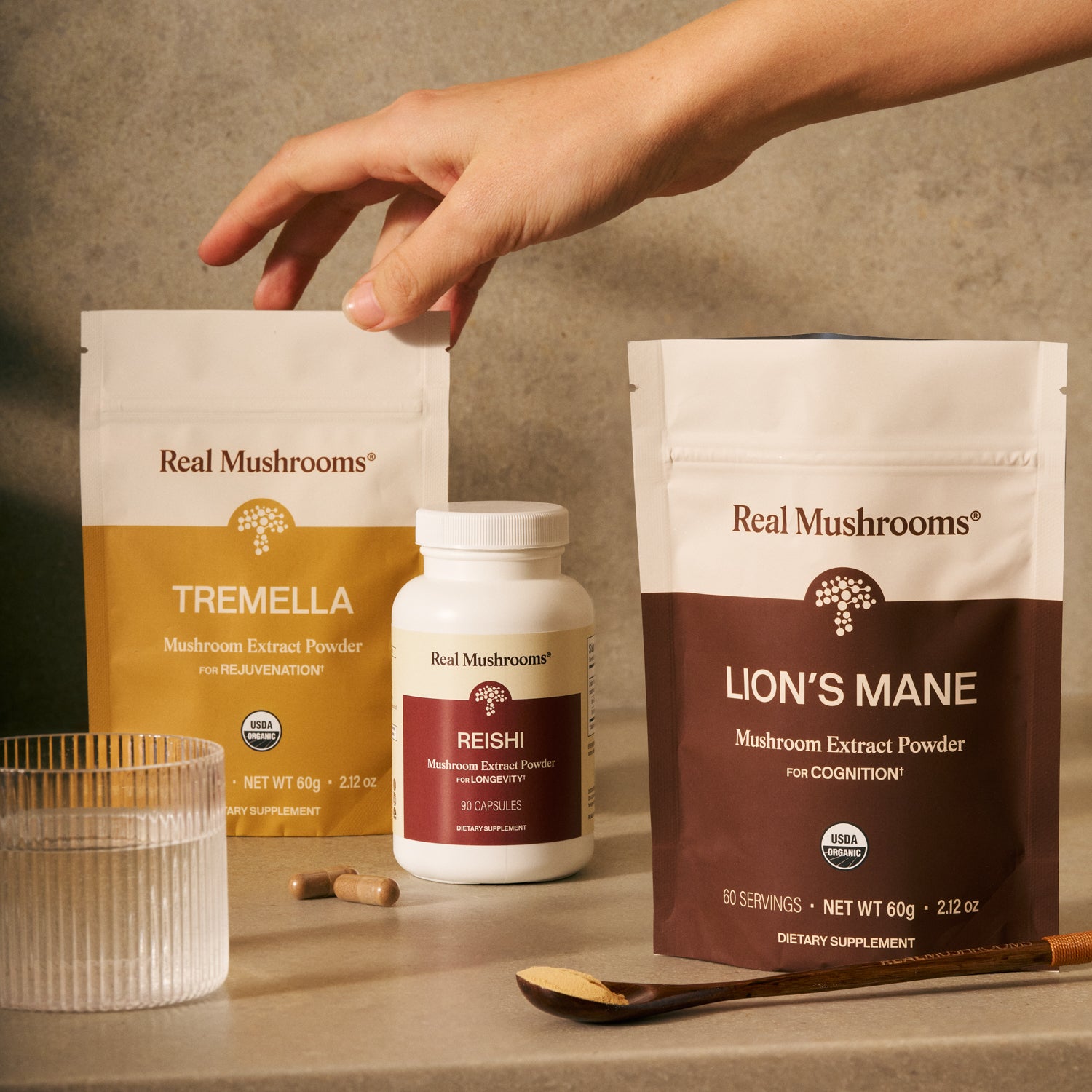 A hand reaches for the Real Mushrooms Longevity Pack—Tremella, Lion's Mane extracts, and Reishi capsules—set beside a glass, capsules, and wooden spoon, all supporting daily brain health.