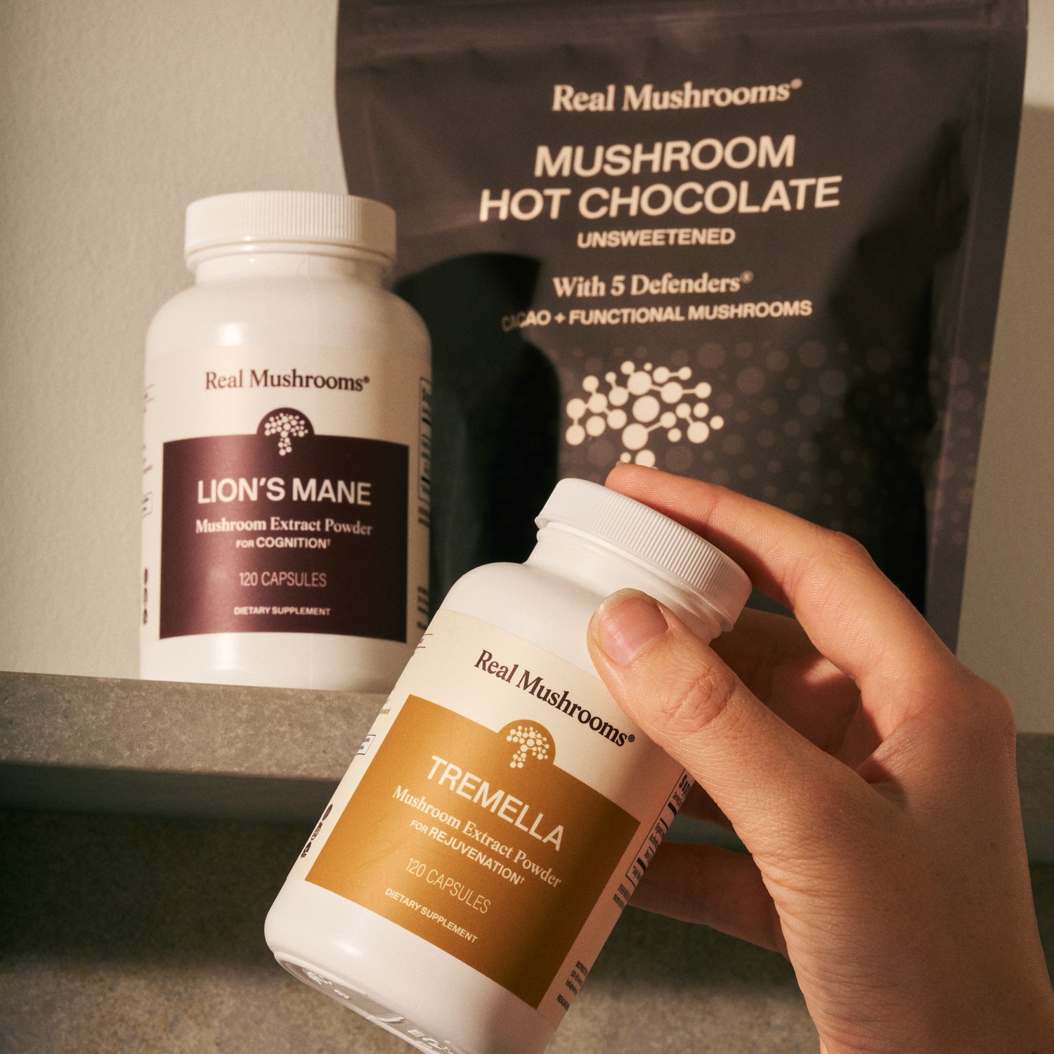 A hand holds a bottle from the Real Mushrooms Feel Good Bundle, with Lion’s Mane capsules and Mushroom Hot Chocolate in the background, all supporting daily wellness.