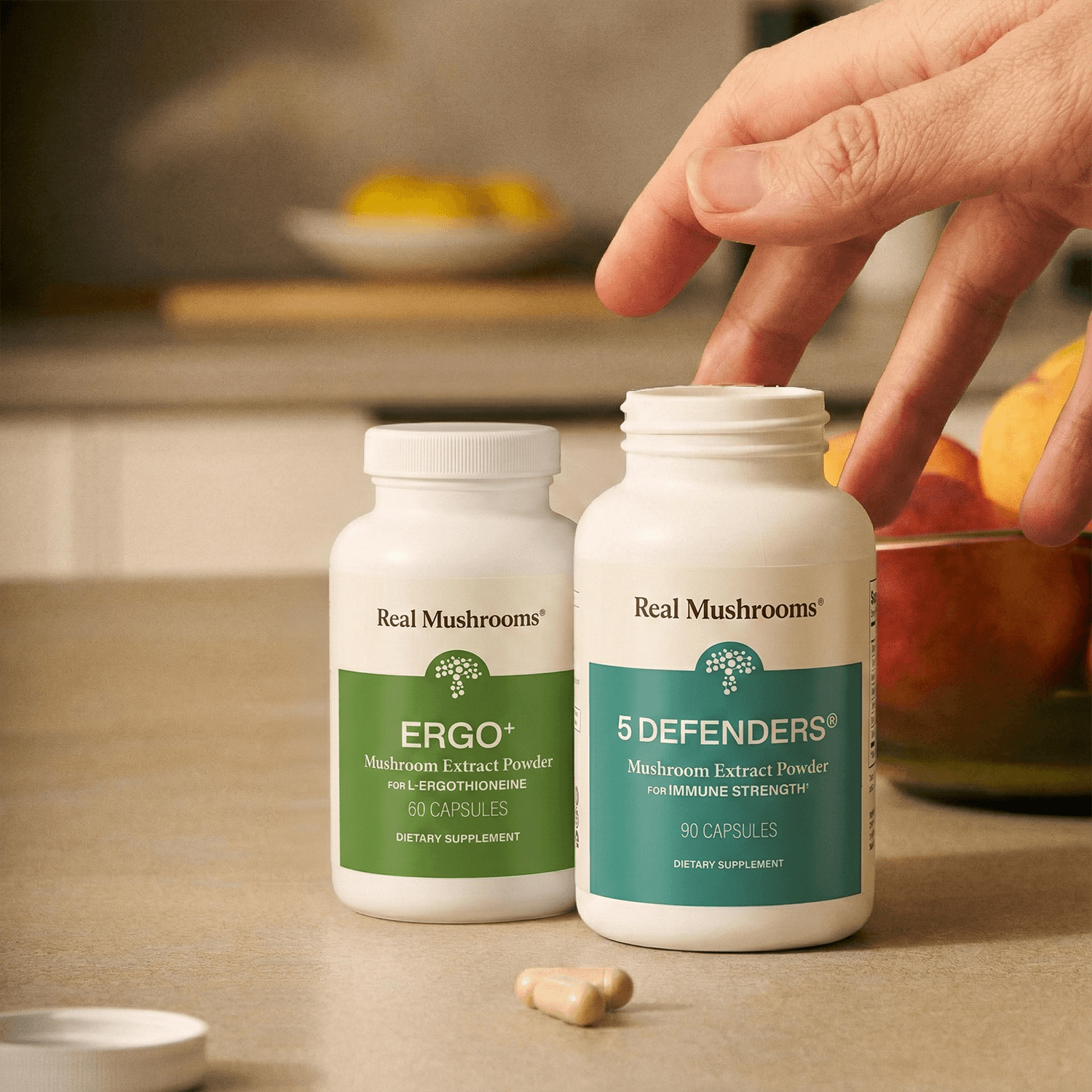 A hand reaches for a white bottle labeled "Immune Power" by Real Mushrooms—a potent mushroom supplement for immune support—next to another bottle on a kitchen counter with capsules and fruit in the background.
