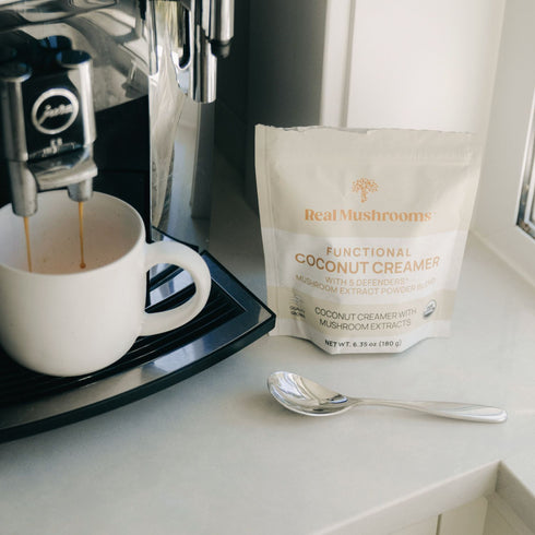 A coffee machine pours espresso into a white mug next to a spoon and a package of Real Mushrooms Functional Coconut Creamer - Powder on the kitchen counter.