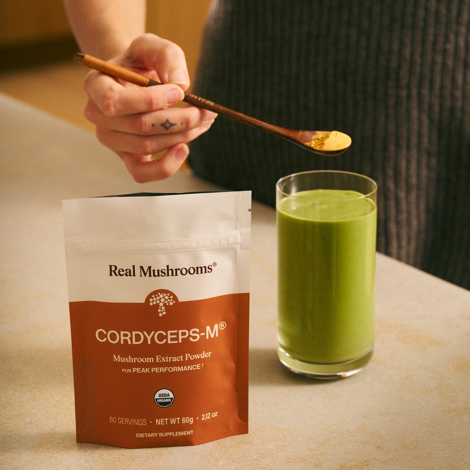 A person adds Real Mushrooms Organic Cordyceps-M® Mushroom Extract Powder – Bulk Supplement from a pouch into a glass of green smoothie with a wooden spoon for natural stamina and energy support.