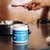 A container of Real Mushrooms Daily Dawg Powder for Pets sits on a countertop, while a person holds a red spoon near a metal bowl in the background.