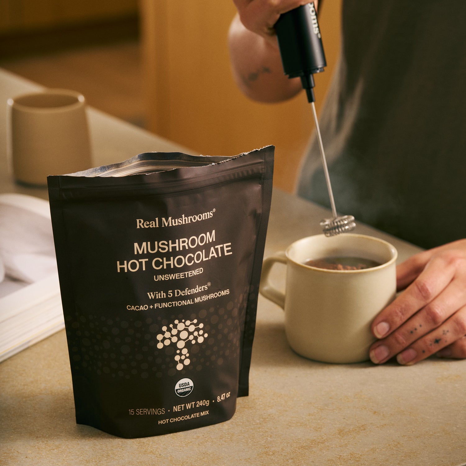Someone uses a milk frother to stir a mug of hot chocolate made with Real Mushrooms’ Mushroom Hot Chocolate Mix, beside a bag of the unsweetened mix on the table.
