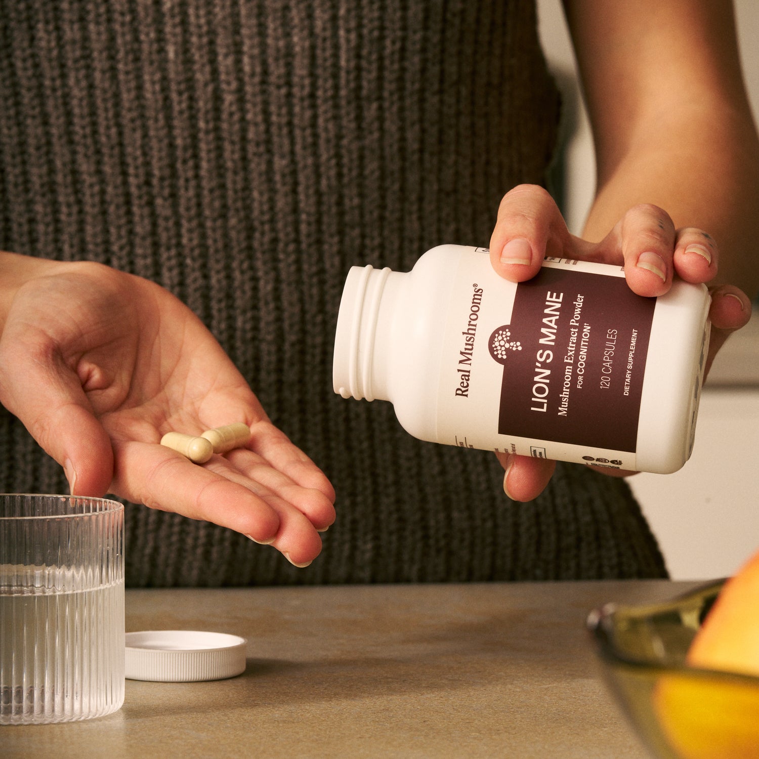 Someone pours two Organic Lion's Mane Extract Capsules by Real Mushrooms into their hand beside a glass of water on the kitchen counter, highlighting support for brain health.