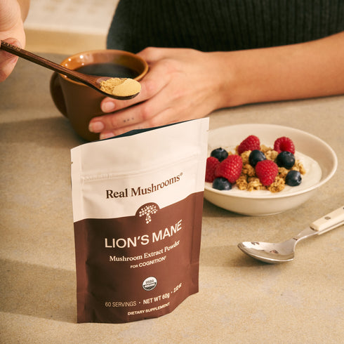 Someone adds Real Mushrooms Lion’s Mane Mushroom Powder, a brain health supplement, to a cup next to a bowl of granola topped with raspberries and blueberries on a table.