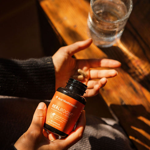 Someone holds a bottle of Real Mushrooms RealBoost—featuring Cordyceps, Guayusa, and Ginseng—in one hand and two capsules in the other, beside a glass of water on a wooden surface.