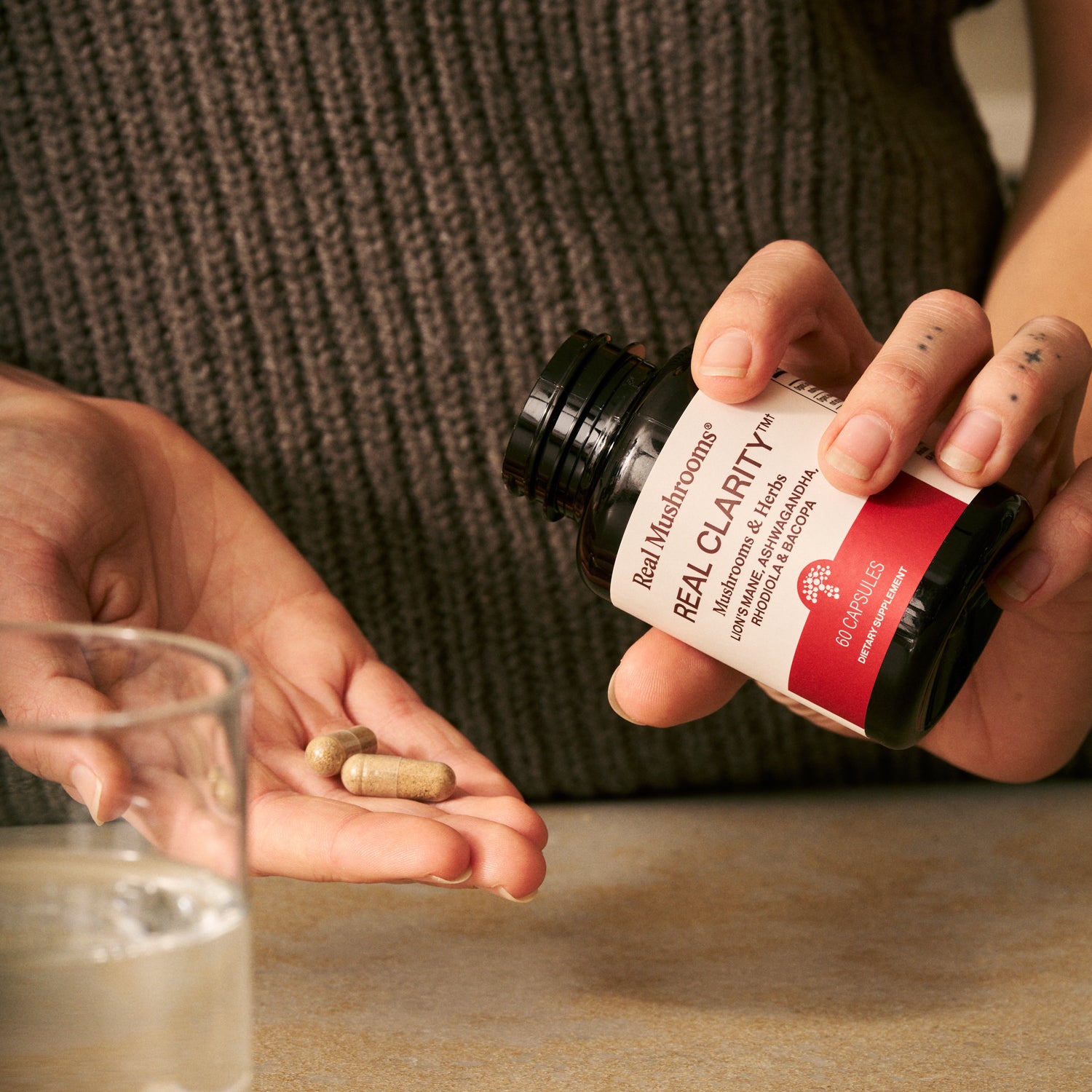 A person pours Real Mushrooms RealClarity capsules, containing Lion's Mane, Ashwagandha, Rhodiola, and Bacopa, into their hand next to a glass of water.