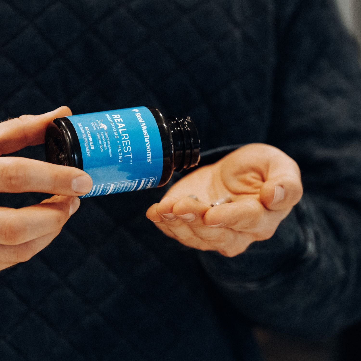 A person pours capsules from a Real Mushrooms RealRest bottle—supplemented with Reishi, Valerian, and Lemon Balm—into their hand.