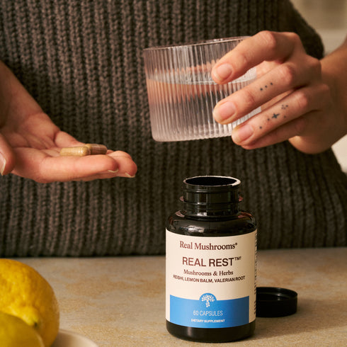 A person holds capsules and a glass of water near a bottle of Real Mushrooms RealRest, which contains calming ingredients like Reishi, Valerian, and Lemon Balm. Lemons are on the table.