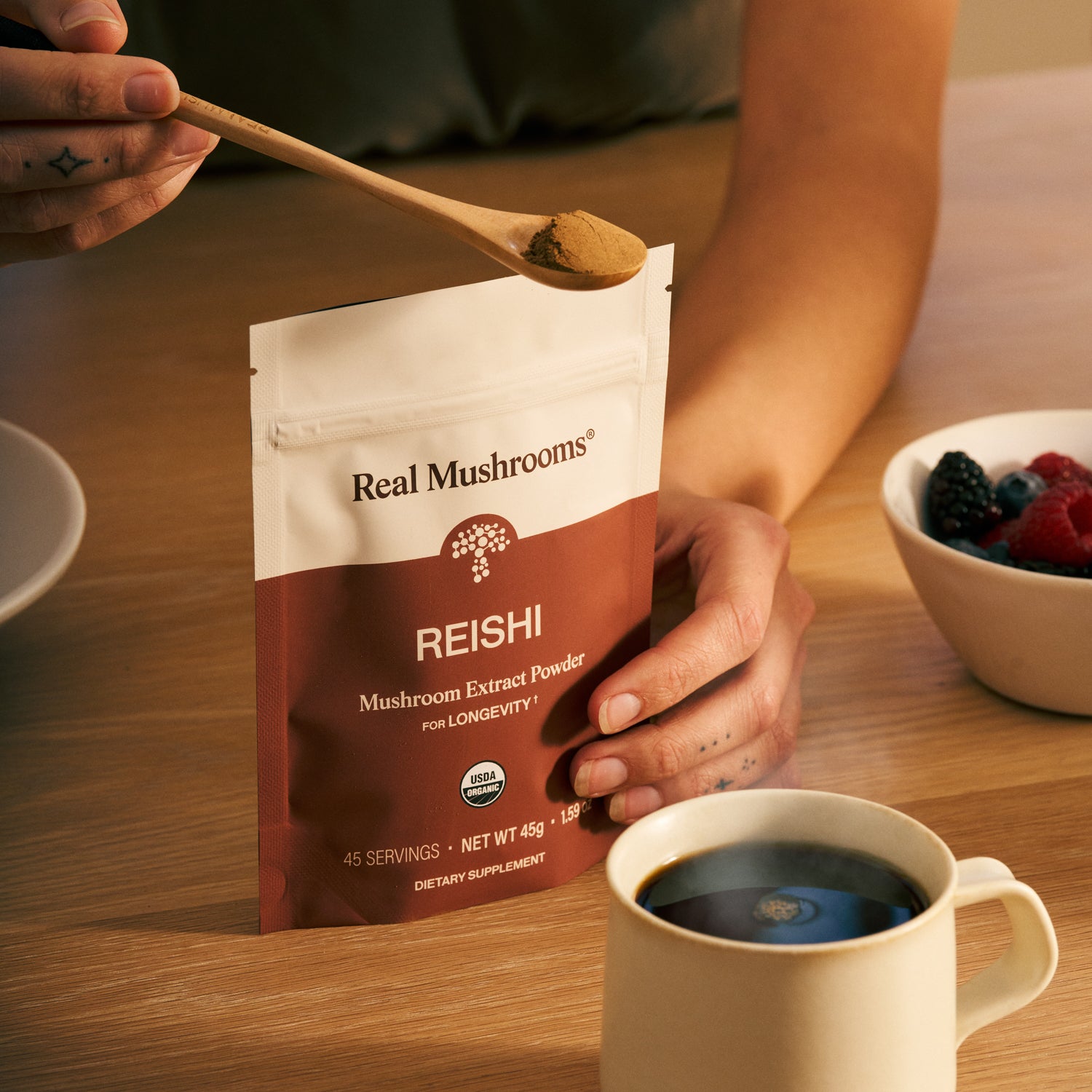 Someone scoops Organic Reishi Mushroom Powder – Bulk Extract from a Real Mushrooms bag, with coffee and berries nearby—showcasing this organic supplement known for its immune-supporting beta-glucans.