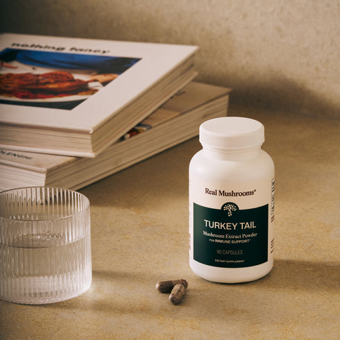 A bottle of Real Mushrooms Turkey Tail Mushroom Capsules, valued for immune-supporting beta-glucans, rests on a countertop beside a glass of water, two capsules, and a stack of books.