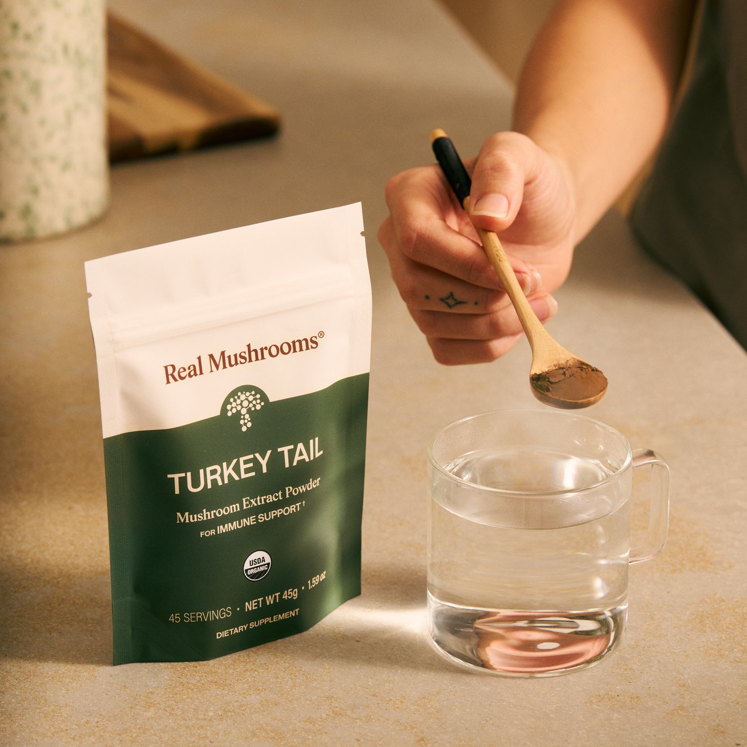 A hand holds a wooden spoon of Turkey Tail Extract - Bulk Powder by Real Mushrooms over a glass of water beside its bag—an organic supplement renowned for supporting immune health.