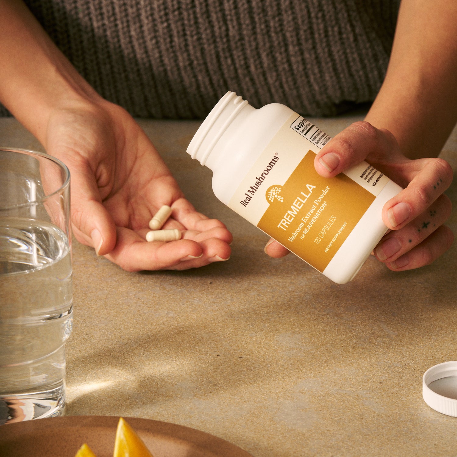 A person pours Organic Tremella Extract Capsules by Real Mushrooms from the bottle into their hand beside a glass of water and lemon wedges on a table.