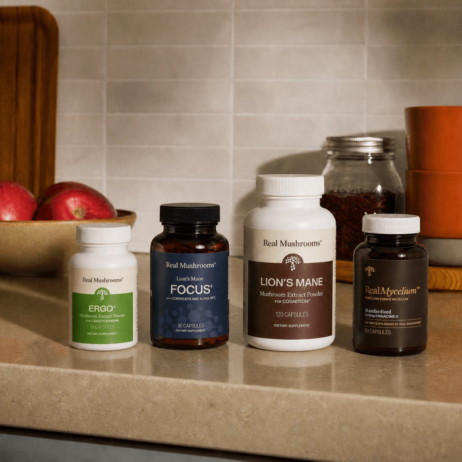 Four bottles from Real Mushrooms’ Brain Boosting Bundle, designed to support brain health and cognitive function, are arranged on a kitchen counter with apples and a ceramic container in the background.
