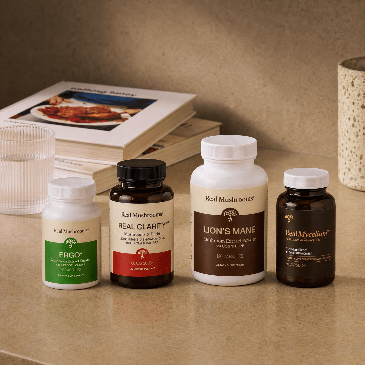Four bottles from the Real Mushrooms Clarity & Vitality Bundle are lined up on a beige countertop, accompanied by a glass of water and books in the background—perfect essentials to support cognitive function and natural energy throughout your day.