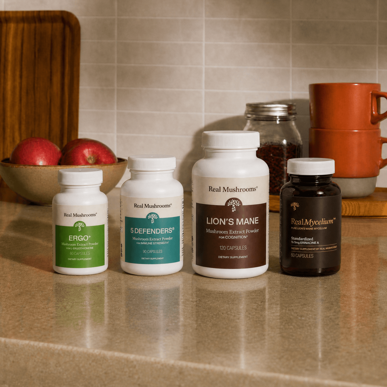 Four bottles of Real Mushrooms’ Complete Mind & Body Bundle are lined up on a kitchen counter, offering powerful mushroom support for brain health. In the background, apples, cups, and jars create a cozy kitchen scene that highlights these supplements from Real Mushrooms, specifically designed to enhance cognitive function.