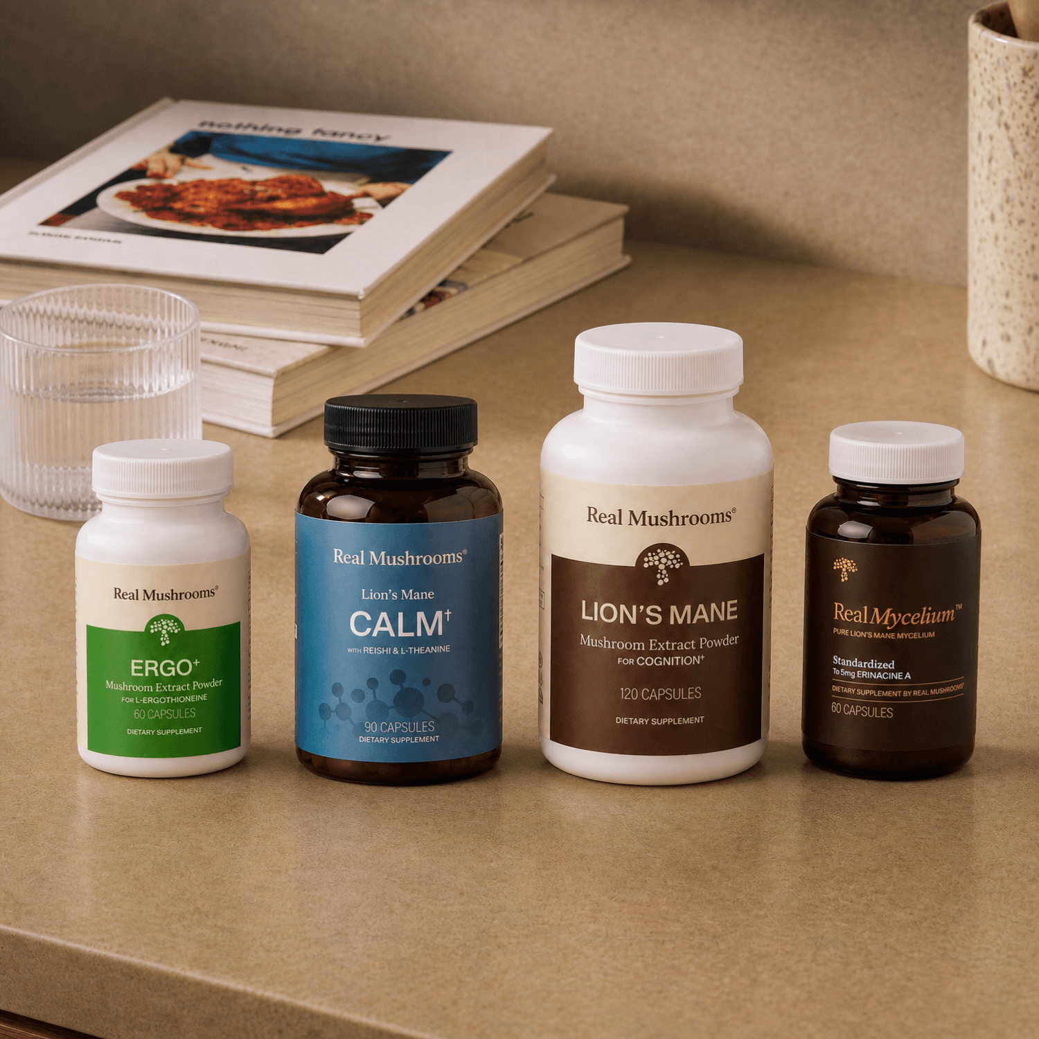 Four mushroom supplement bottles from Real Mushrooms—ERGO, Lion’s Mane CALM, Lion’s Mane, and RealMycelium—sit on a kitchen counter in front of two cookbooks, showcasing the Rest & Restore Bundle by Real Mushrooms: the perfect addition to your daily routine for supporting brain health and mood.
