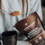 A person holds a spoonful of USDA Certified Organic Hot Chocolate Sample powder above a black mug, with a brown packet labeled "Real Mushrooms" nearby.