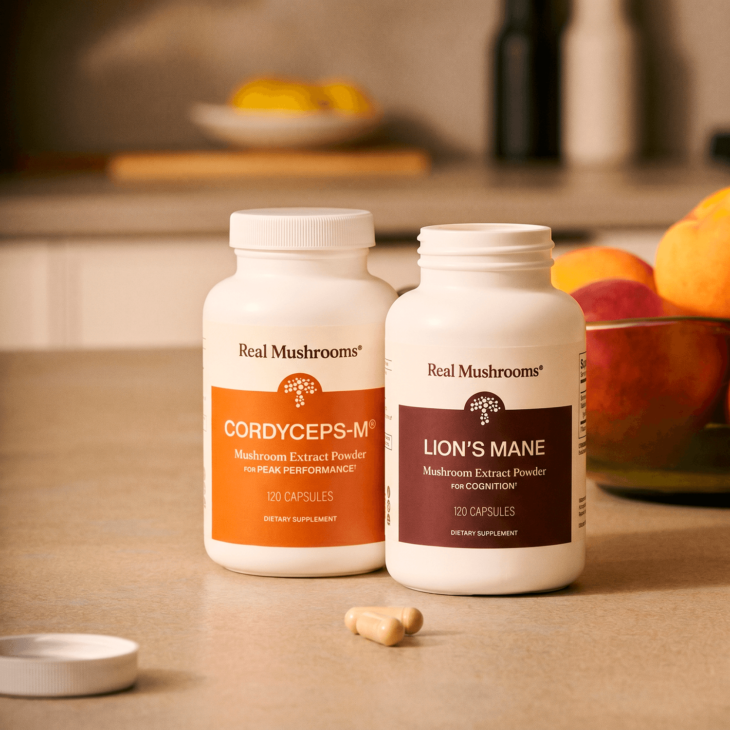 Two bottles of Real Mushrooms Energy Duo supplements and two capsules sit on a kitchen counter, showcasing premium mushroom extracts; a bowl of peaches is in the background.