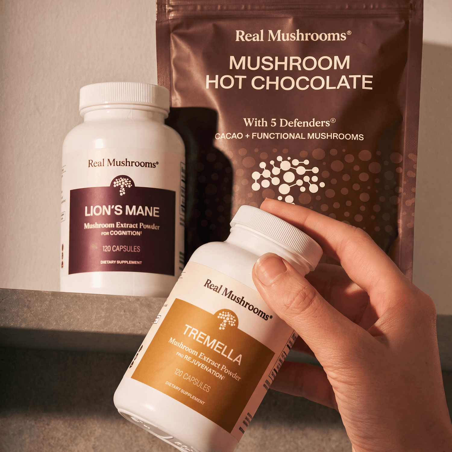 A hand holds a bottle from the Feel Good Bundle by Real Mushrooms, featuring Lion’s Mane capsules and hot chocolate powder—all expertly crafted with organic mushroom extracts—displayed together on a shelf.