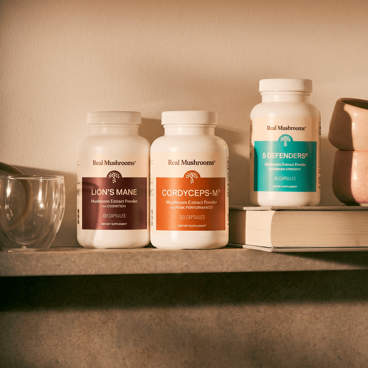 Three bottles of Real Mushrooms’ The Big 3, featuring potent mushroom extracts for brain and immune support, are arranged on a shelf beside a glass, stacked books, and a ceramic cup in soft, warm lighting.