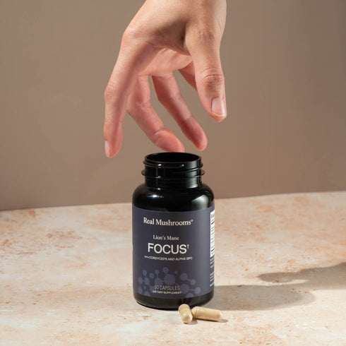 A hand reaches for a black bottle labeled "Real Mushrooms Focus Power Trio," emphasizing its brain health benefits, with two capsules resting on a beige surface nearby.