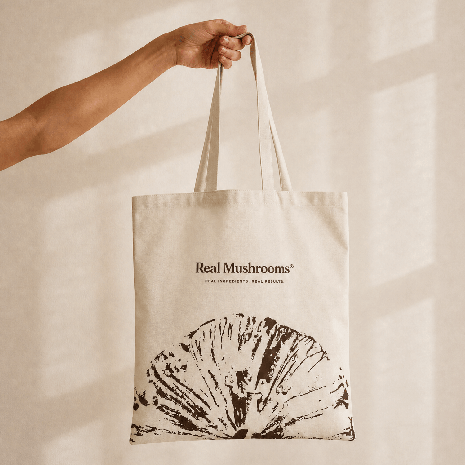 A hand holds the Real Mushrooms Tote Bag, featuring a large brown mushroom illustration and “Real Mushrooms” text printed on the front.