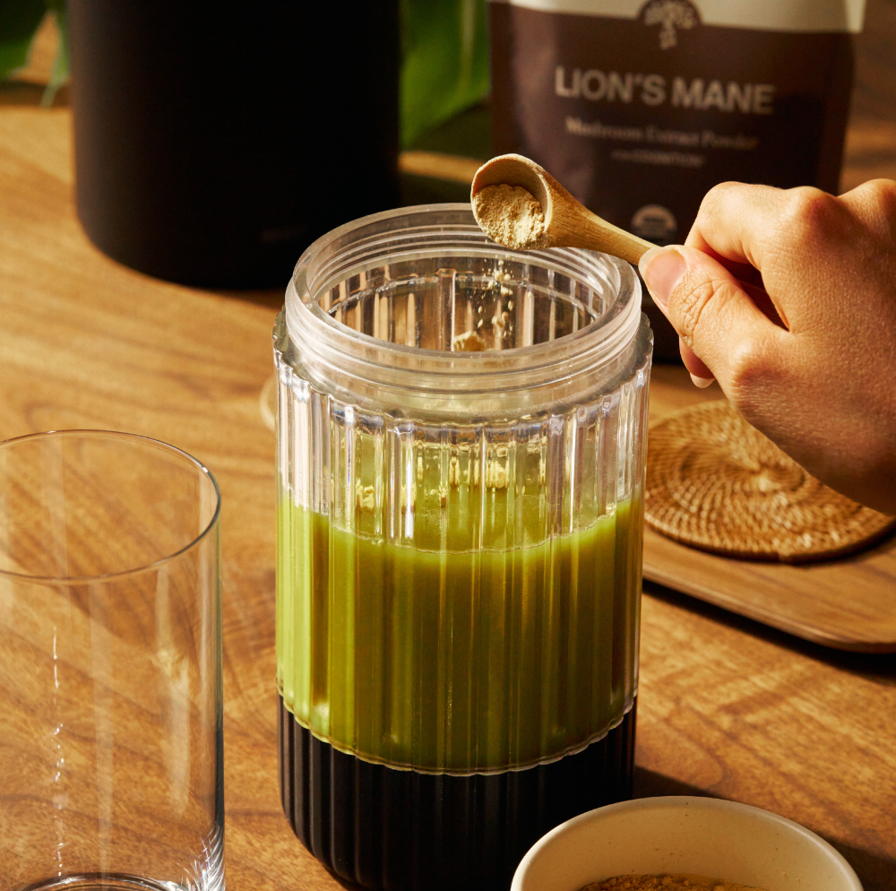 A hand adds powdered supplement to a glass of green liquid on a wooden table, with a packet labeled "Lion's Mane" in the background.