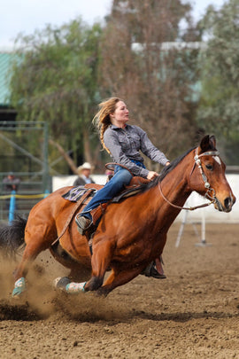 A woman wearing jeans and a long-sleeve shirt rides a brown horse at speed on a dirt arena, kicking up dust as she turns.