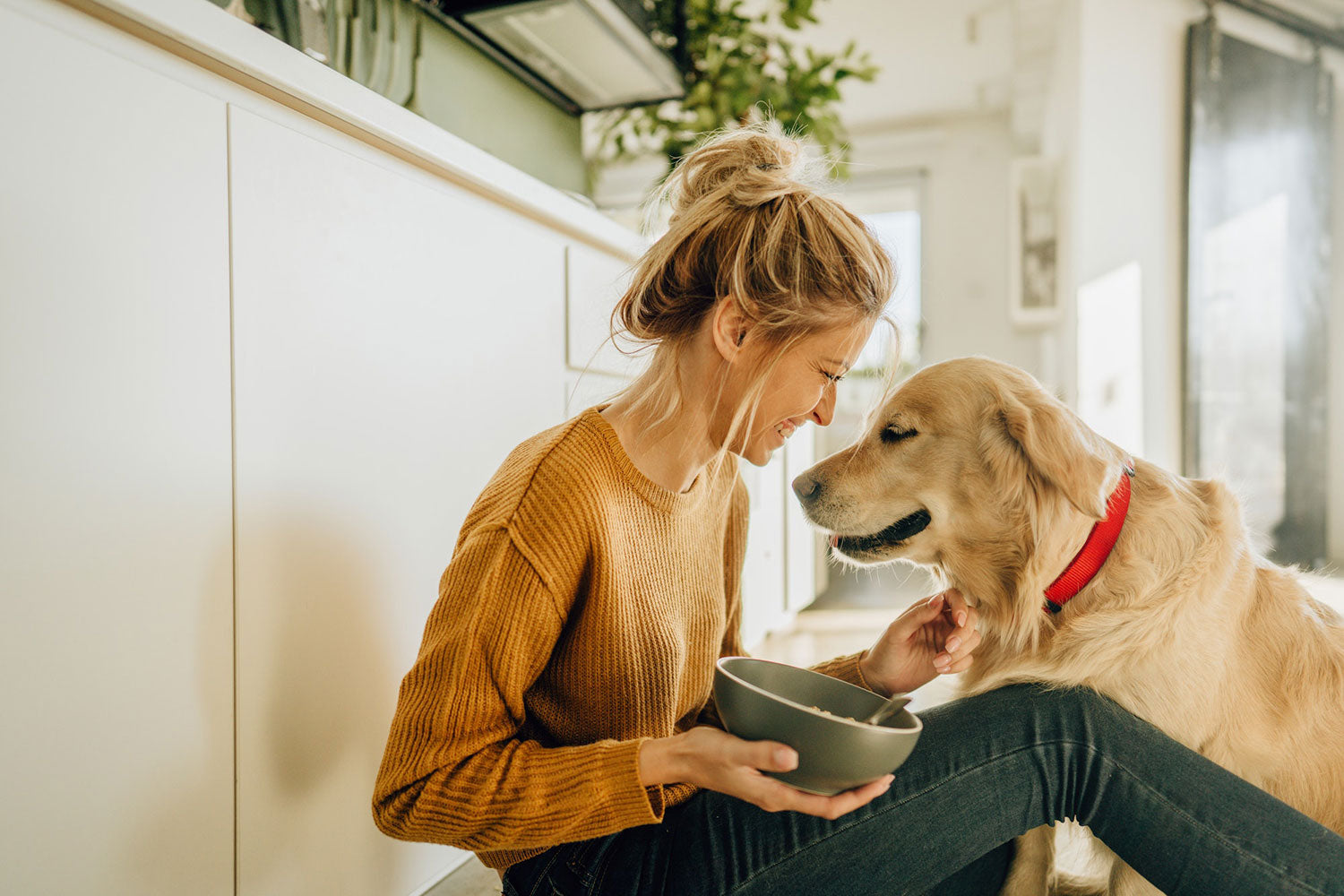 Woman sitting on the floor with a dog, both looking at a bowl in a kitchen.