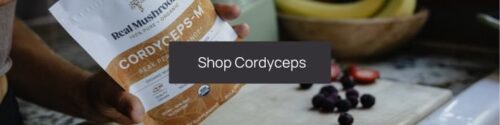 A person holding a packet labeled "Caterpillars with Cordyceps" in a kitchen with fruits in the background and a "shop cordyceps" clickable banner overlay.