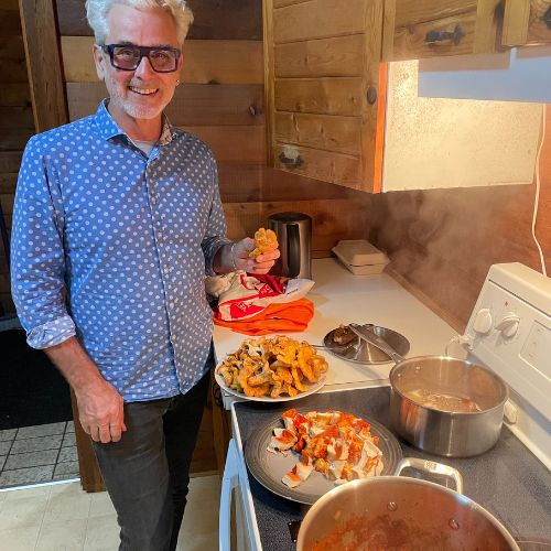 Man smiling in a kitchen holds mushrooms, with more on a plate and a pot cooking on the stove, perfecting his lobster mushroom recipe.