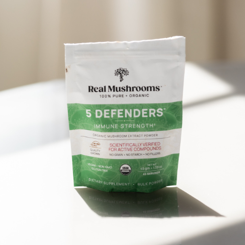 Bag of "5 Defenders" organic mushroom extract powder by Real Mushrooms sits on a reflective surface. Packaging highlights immune strength, scientific verification, and the benefits of mushroom vitamins.