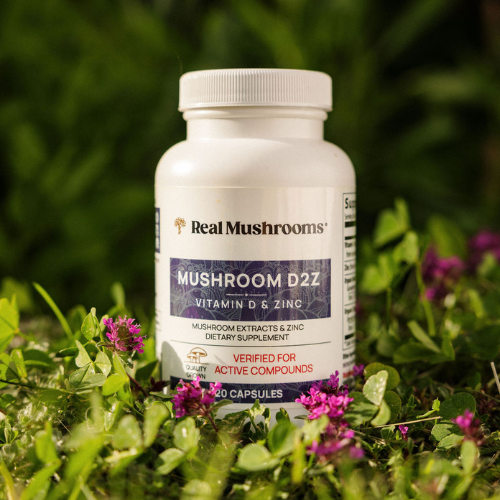 A bottle of Real Mushrooms Mushroom D2Z dietary supplement, rich in mushroom vitamins, vitamin D, and zinc, is nestled among green foliage and small purple flowers.