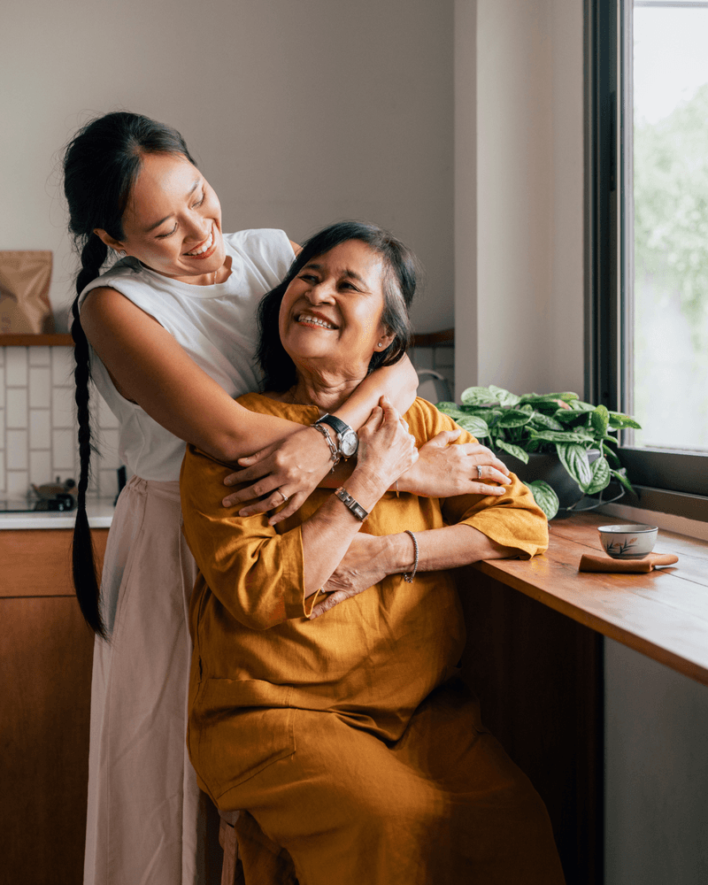 A woman hugging an older woman (mother) sitting by the window